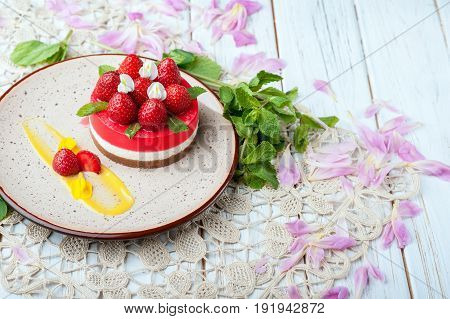 Mini cake layered with cream and red strawberries on top on beautiful plate decorated with leaves of mint flowers and staying on napkin. Wooden white background. Food dessert healthy concepts.