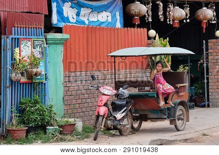 Cambodia, Siem Reap 12/08/2018 A Little Asian Girl Sits In A Moto Rickshaw Near A House With Red Lan