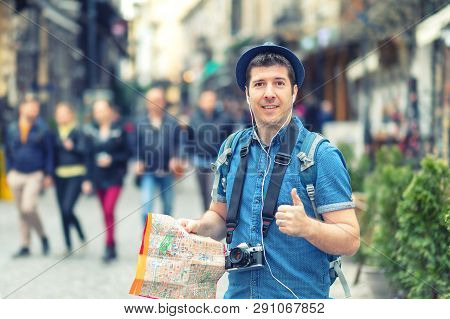 Traveler With Trendy Look Holding Map Showing Thumb Up On Street Full Of Restaurants And Shops Of Eu