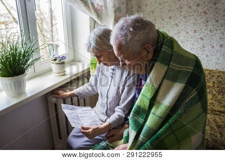 The Senior Woman Holding Gas Bill In Front Of Heating Radiator. Payment For Heating In Winter.
