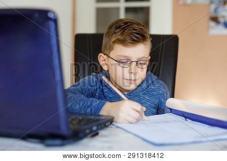 Happy Healthy Kid Boy With Glasses Making School Homework At Home With Notebook. Interested Child Wr