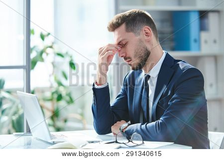 Exhausted businessman in formalwear sitting by desk in front of laptop after hard working day