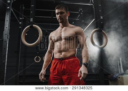 Portrait Of Young Muscular Athlete Preparing For Workout At The Gym. Cloud Of Dust Chalk Powder On D