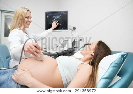 Pretty Pregnant Woman Lying In Medical Cabinet In Modern Clinic On Consultation. Female Doctor, Blon
