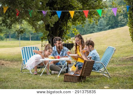 Dedicated young parents of two children listening to their funny son talking while eating together during family picnic in a summer day