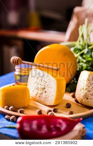 Cheese Wheel On A Wooden Board Lies On The Table With Food And Fresh Herbs. Blurred Background.