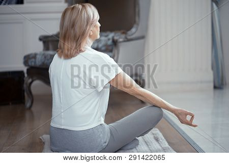 Woman Meditates At Home Looking Out The Window. She Sits On The Floor In An Asana Position. The Woma