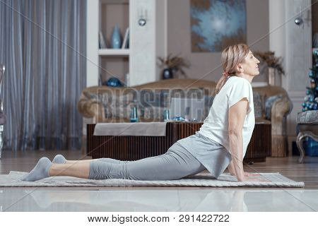 Yoga Classes For Women Over 50 Years. A Woman Is Engaged At Home In The Living Room. On The Carpet S