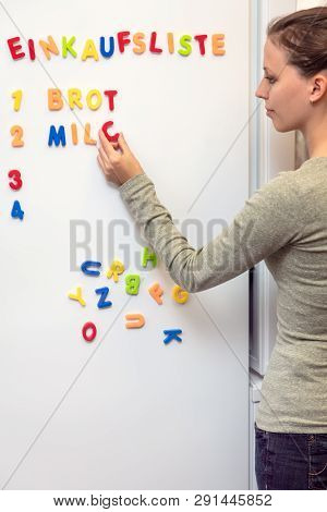 Young Woman Notes The Shopping List On The Fridge With Colorful Magnet Letters, German Word Einkaufs