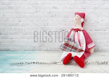 Tilda Doll In A Red Cap, And In A Red Dress With Four-piece. The Doll Sits On A Wooden Light Table. 