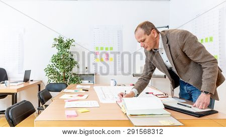 Senior production manager going over design drawings for final approval and revisions in a large, brightly lit office with production planning boards on the walls.