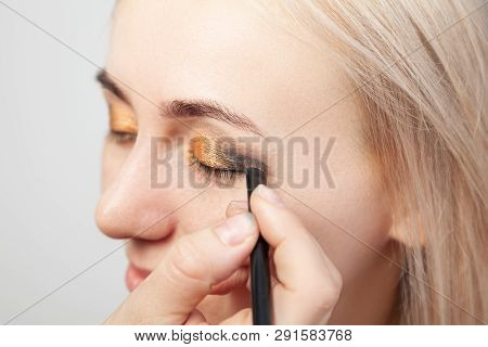 Visagiste With A Brush In One Hand Putting Makeup On The Eyes Of The Model, The Other Hand Pulls The
