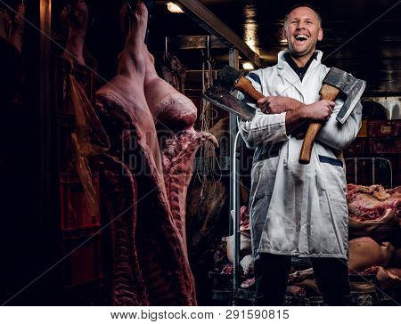 Cheerful Butcher In Workwear Posing With Two Axes In A Refrigerated Warehouse In The Midst Of Meat C