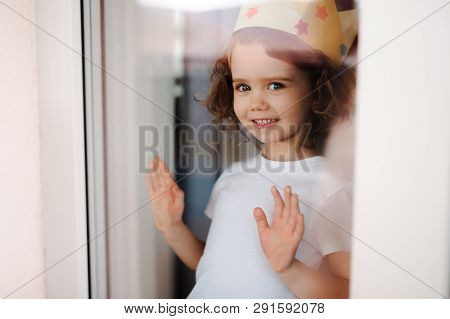 Portrait Of Small Girl With Crown At Home, Looking Out Of A Window. Shot Through Glass.