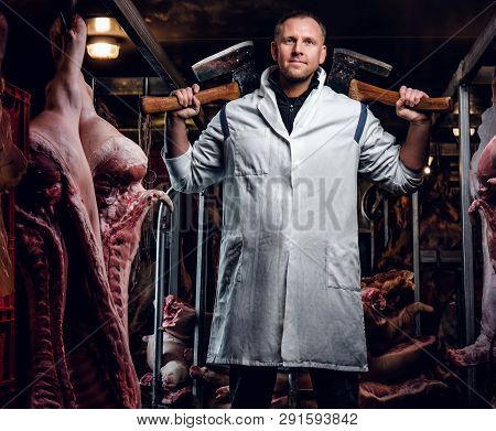 The Butcher In Workwear Posing With Two Axes In A Refrigerated Warehouse In The Midst Of Meat Carcas