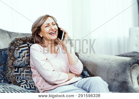 An Elderly Woman Talking On The Phone. Woman Sitting At Home On The Couch. She Has Fun She Laughs Wi