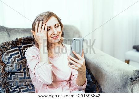 Adult Woman Resting At Home In The Living Room. She Has A Phone In Her Hands. She Gently Smiles And 