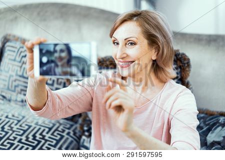 Elderly Woman At Home In The Living Room With A Phone In Her Hands. A Woman Wants To Take A Picture 