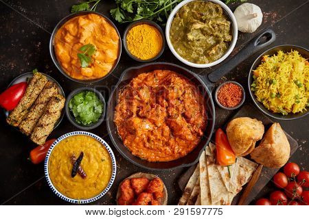 Assortment of various kinds of Indian cousine on dark rusty table. Chicken Tikka Masala, Butter, Nilgiri, Daal Tarka. Served with fried rice, naan bread and spices.