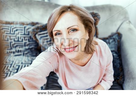 An Adult Woman With A Smile On Her Face Makes A Selfie. A Woman Is Sitting On The Sofa At Home. In A