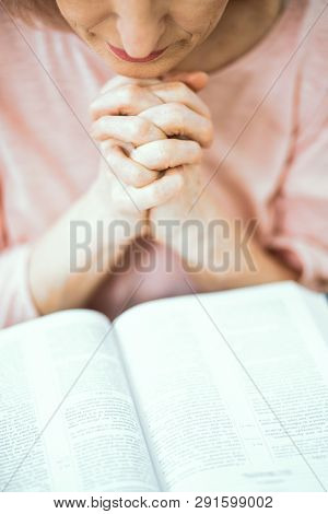 An Elderly Woman Prays With Folded Hands. Before It Lies The Book Of The Bible. Close Up Shot.