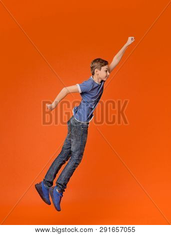 Little Boy Simulating Flight, Jumping With Raised Fist Like Superhero On Orange Background