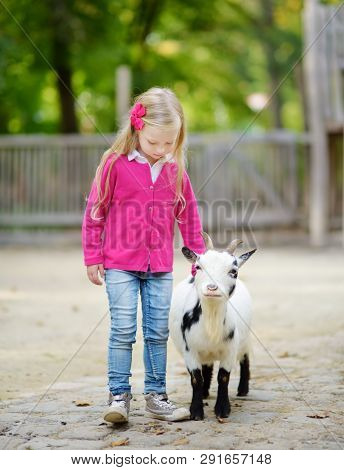 Cute Little Girl Petting And Feeding A Goat At Petting Zoo. Child Playing With A Farm Animal On Sunn