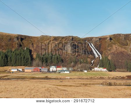 Waterfall And Village In South Iceland, With River And Fields In The Foreground