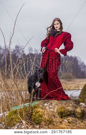 Art Photography. Beautiful Fairy Princess In Red Dress With Crown And Her Dog On Leash In Forest On 