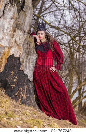 Art Photography. Beautiful Fairy Princess In Red Dress And Spiky Black Crown In Forest In Early Spri
