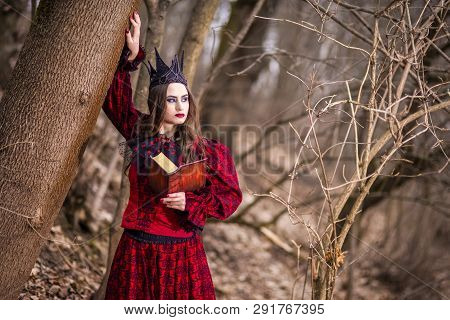 Art Photography. Gorgeous Mysterious Fairy Princess In Red Dress And Black Crown With Old Book. Posi