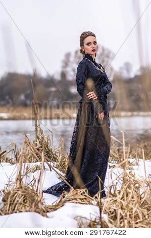 Young Caucasian Brunette Female In Art Dress With Necklace And Crown Posing Against River Outdoors. 
