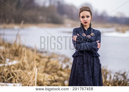 Passionate Caucasian Brunette Female In Art Dress With Necklace And Crown Posing Against River Outdo