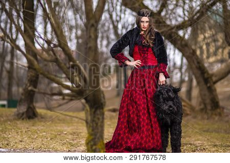Beautiful Princess In Red Dress And Black Fur Jacket Posing In Crown Along With Her Dog In Forest Du