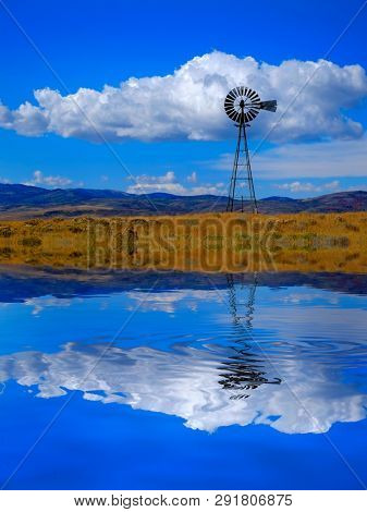 Windmill on hillside in countryside rural America with sky and clouds reflection in water like a lke or pond reflect mirror