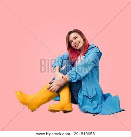 Smiling Young Girl Sits On Floor Wearing Spring Raincoat Protected From The Rain And Tall Yellow Rub