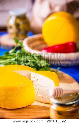 Cheese Wheel On A Wooden Board Lies On The Table With Food And Fresh Herbs. Blurred Background.