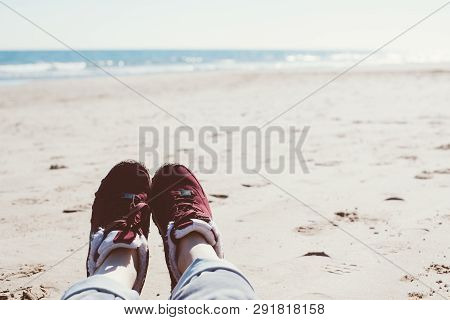 Feet Of Young Woman In Sport Footwear And Jeans Relaxing After Hiking On Sand Beach. Pov. Adventure 