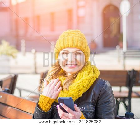 Portrait Of A Young Attractive Woman In A Knitted Hat And Knitted Scarf, Which Sits On A Bench In Th