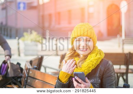 Portrait Of A Young Attractive Woman In A Knitted Hat And Knitted Scarf, Which Sits On A Bench In Th