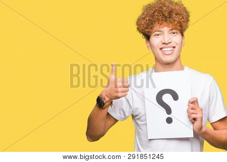 Young handsome man holding paper with question mark happy with big smile doing ok sign, thumb up with fingers, excellent sign