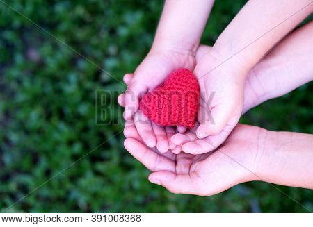 Red Knitted Heart In The Hands Of A Child And Mother On A Green Background. The Concept Of Love, Mer
