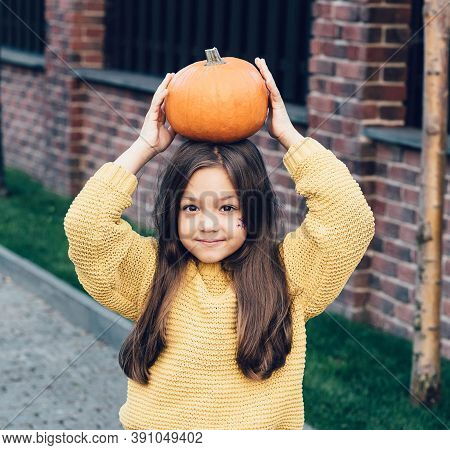 Funny Child Girl In Orange Pullover For Halloween With Pumpkin And On A Dark Brick Background.