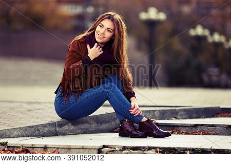 Happy young fashion woman sitting on street sidewalk  Stylish female model in red leather fringe suede jacket and dark blue jeans