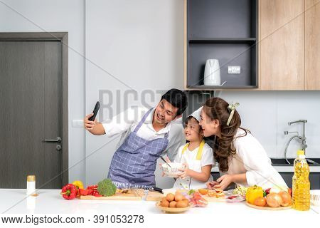 Young Asian Family Are Preparing Salad In The Kitchen And Father Take A Photo Selfie By Phone. Excit