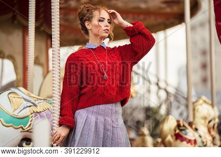 Pretty Teenager Redheared Girl Is Wearing Red Sweater Staying Near Colorful Carousel , Autumn Time