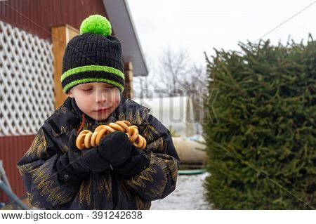 A European Boy In A Warm Jacket And Hat Stands In Winter Near A Fir Tree. The Child Holds The Steeri