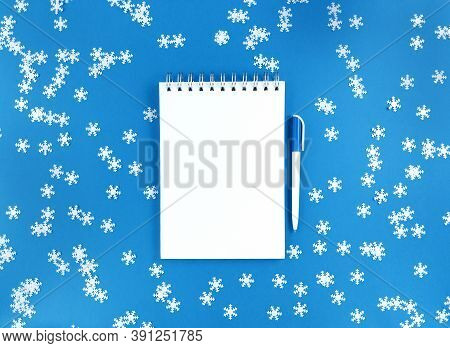 White Blank Sheet Of Notebook And A Pen On A Blue Background With Scattered Confetti Snowflakes. Hol