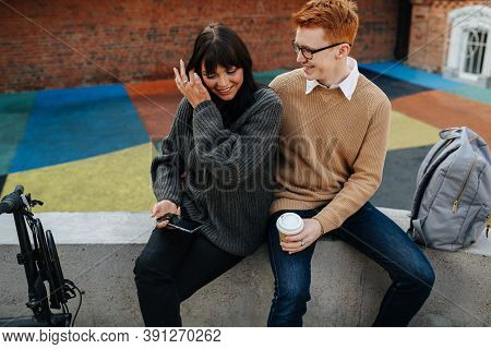 Two Happy People In Love Sitting On A Concrete Border, Looking At Phone