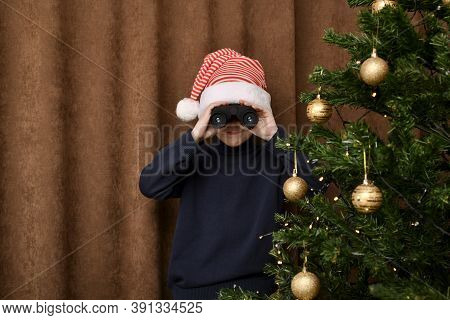 A Boy With Compressed Lips, Impatiently, In A Striped Cap, Looks Out Through The Binoculars Of Santa
